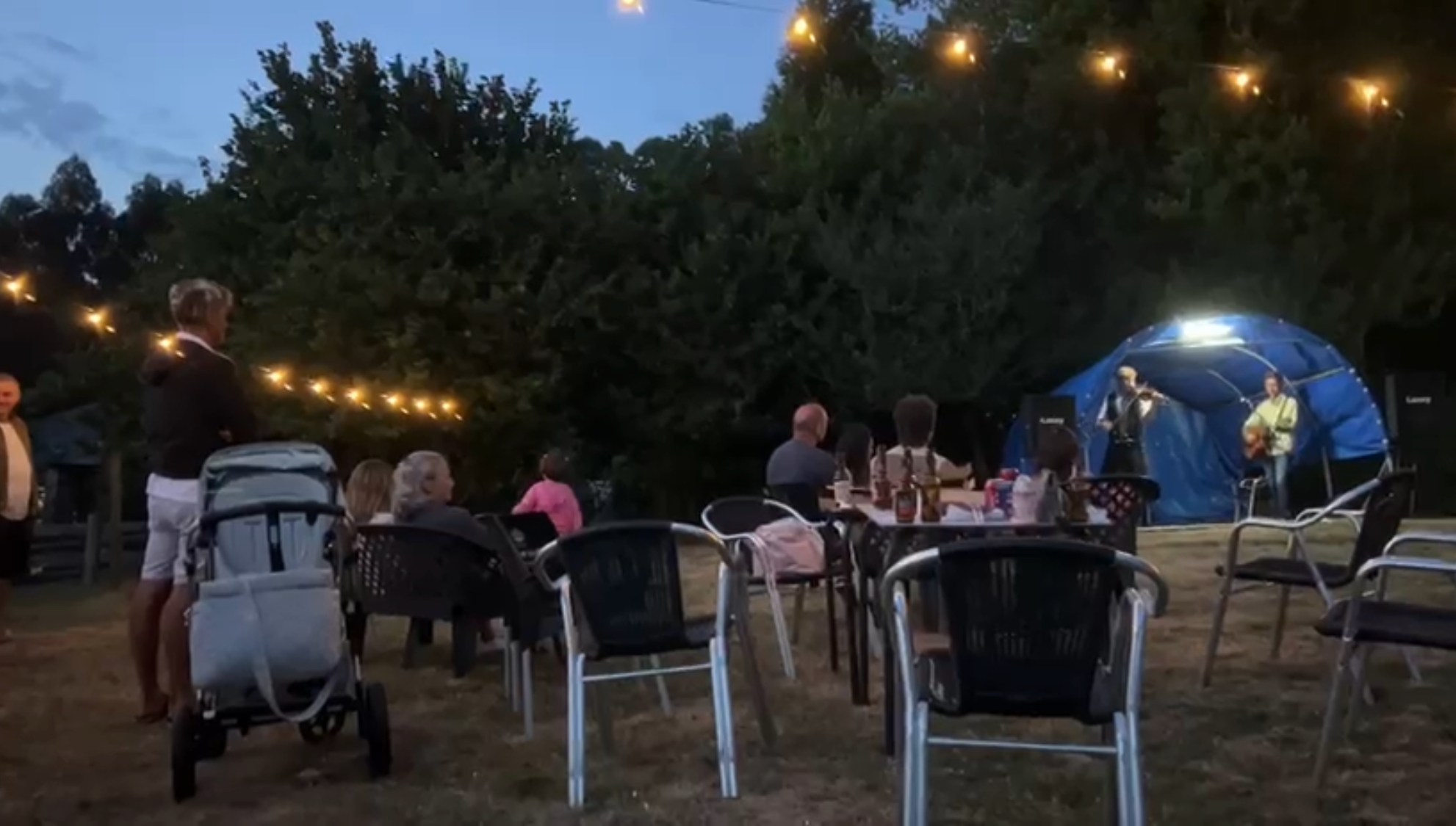 Un concierto al anochecer en Lamacido, con público sentado, guirnaldas de luces y músicos bajo una cubierta azul.